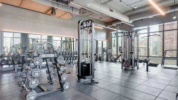 A gym with various weightlifting equipment, including dumbbells and weight plates, and large windows offering views of the city.