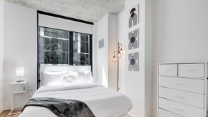Un dormitorio moderno con un ventanal grande, una lámpara de noche, una cama con cabecero blanco y un aparador blanco.