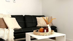 A modern living room with a black leather sofa, a white coffee table with a woven basket, a wall clock, and floating shelves with books and pumpkins. - Beautiful Park Side Apartment (Steinbach)