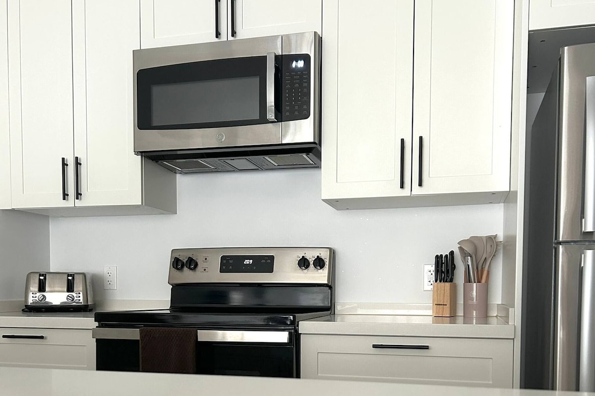 A modern kitchen with white cabinets, a stainless steel microwave, a toaster, an oven, and a refrigerator.