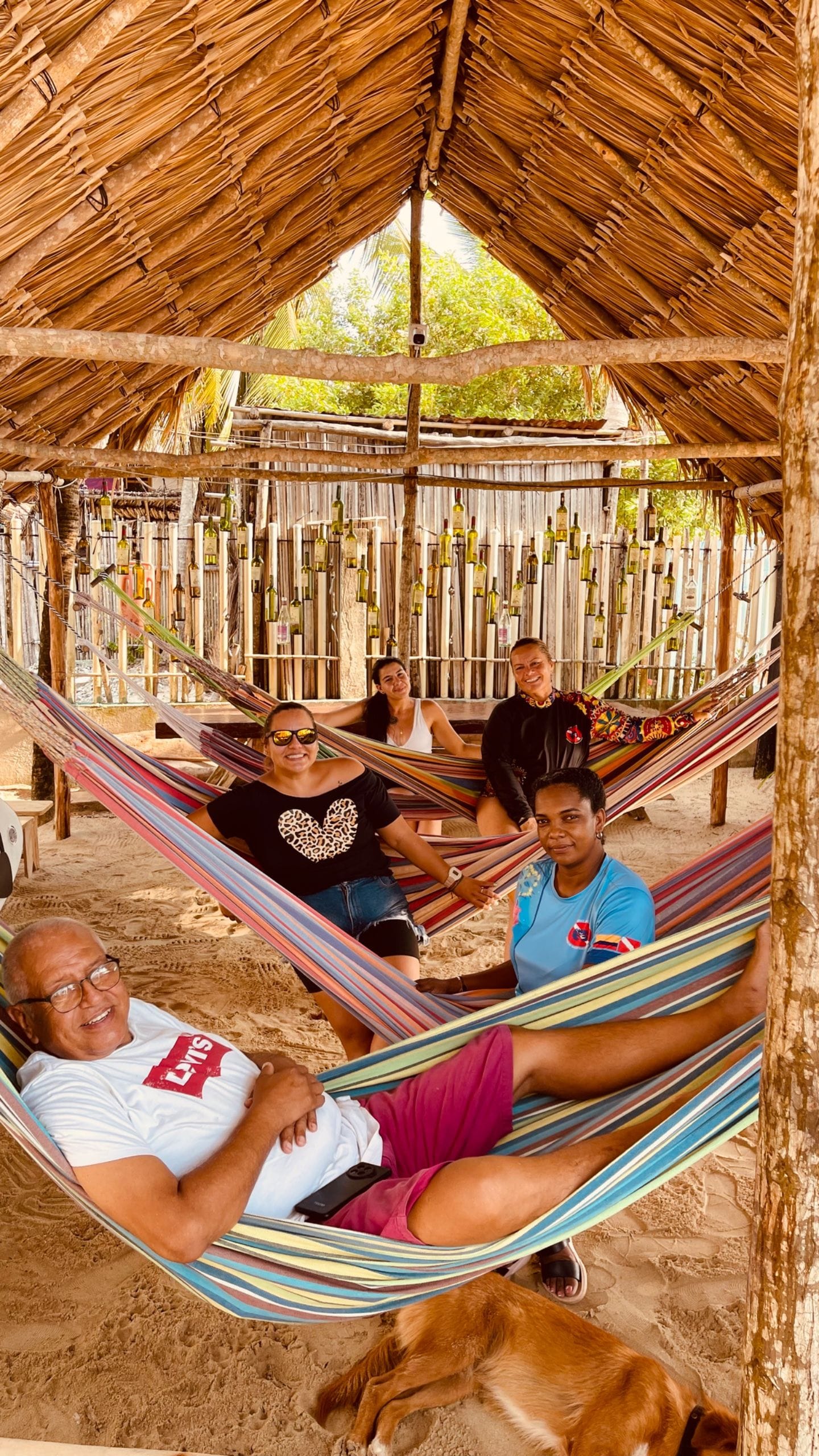 Un grupo de personas descansando en una hamaca bajo una estructura de techo de paja, con botellas colgadas en la pared detrás.
