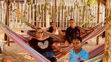 Un grupo de personas descansando en una hamaca bajo una estructura de techo de paja, con botellas colgadas en la pared detrás.