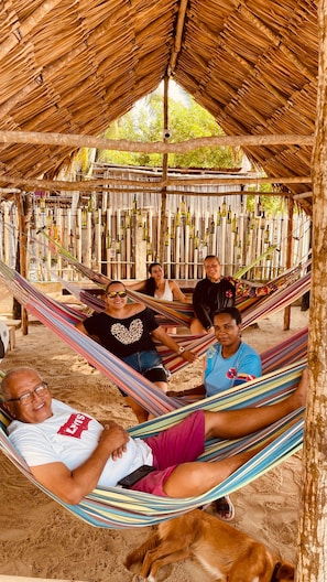 Un grupo de personas descansando en una hamaca bajo una estructura de techo de paja, con botellas colgadas en la pared detrás.