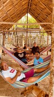 Un grupo de personas descansando en una hamaca bajo una estructura de techo de paja, con botellas colgadas en la pared detrás.