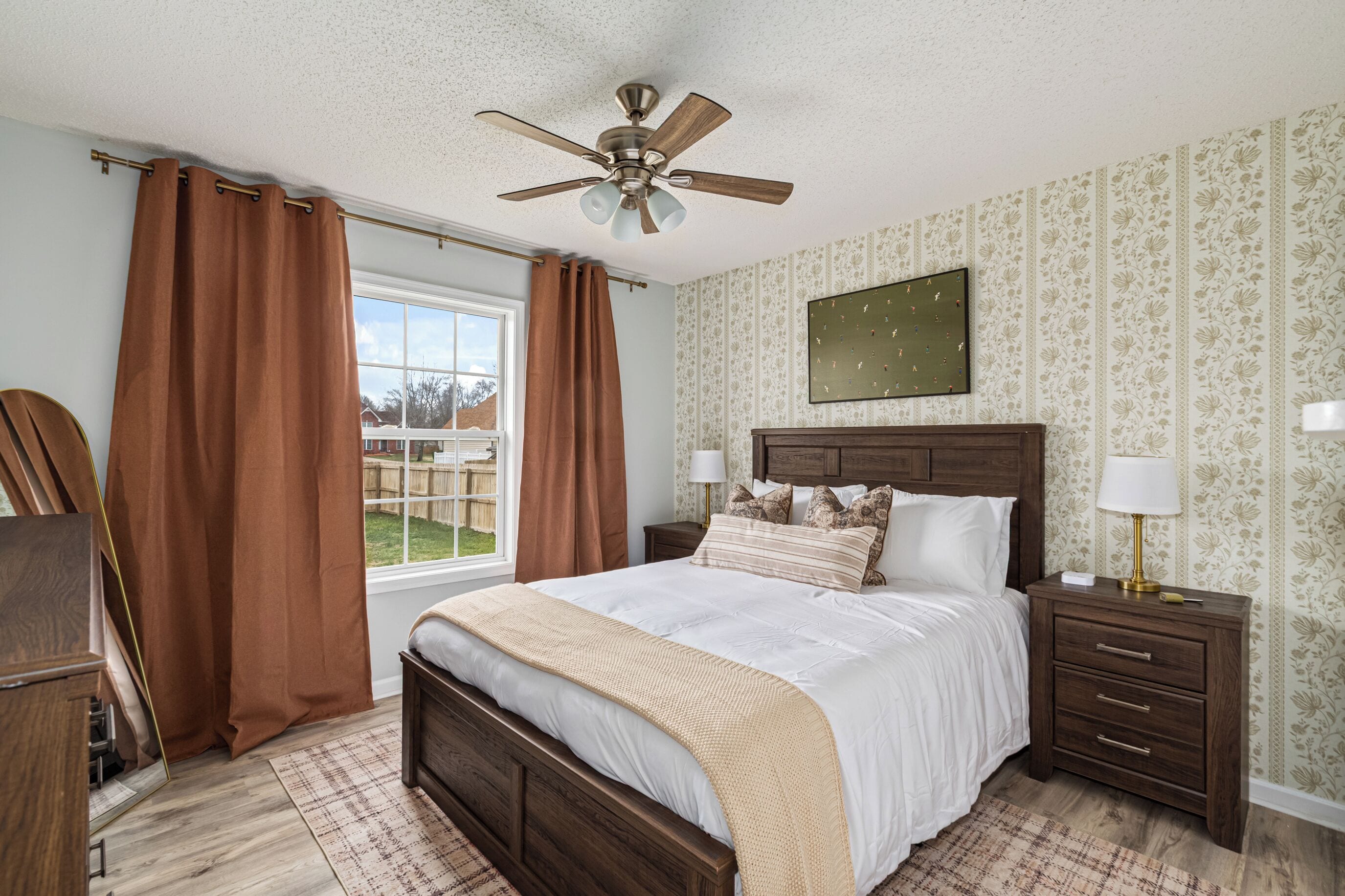 A bedroom with a wooden bed, two bedside tables with lamps, a ceiling fan, and a window with curtains.