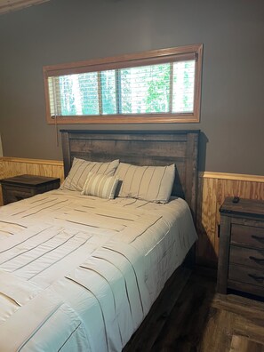 Quarto com cama grande, cabeceira de madeira, janela com persianas e mesa de cabeceira com luminária.