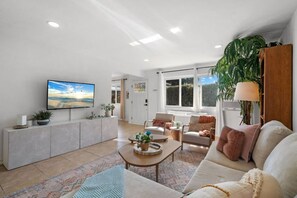 A spacious living room with a large flat-screen TV, comfortable seating, a coffee table, and a large window providing natural light. - Boho Palms by Coastline Vacation Rentals (San Diego)