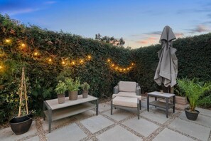 An outdoor patio with a sofa, armchair, and table, surrounded by potted plants and a string of lights. - Boho Palms by Coastline Vacation Rentals (San Diego)