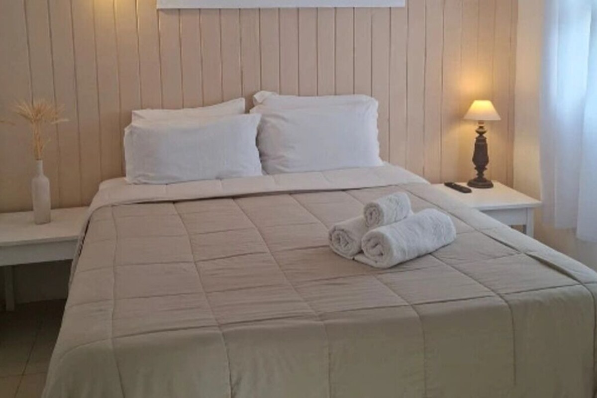 A neatly made bed with a beige bedspread, two pillows, and rolled towels on the side. There is a bedside table with a lamp and a vase of dried plants.