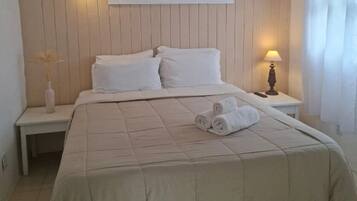Una cama bien hecha con una colcha color beige, dos almohadas y toallas enrolladas al lado. Hay una mesita de noche con una lámpara y un florero con plantas secas.