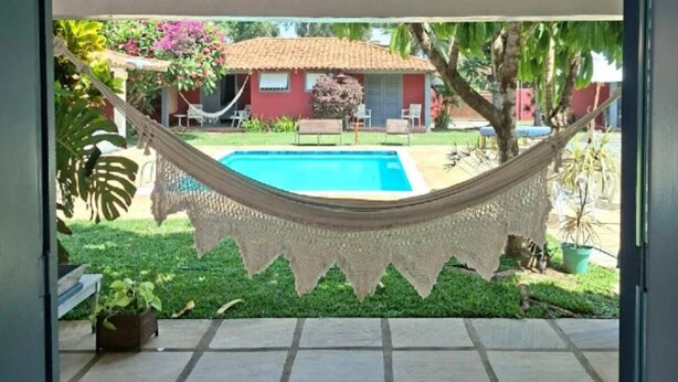 A hammock over a swimming pool with a house and garden in the background. - POUSADA DOS PÁSSAROS (Rio de Janeiro)