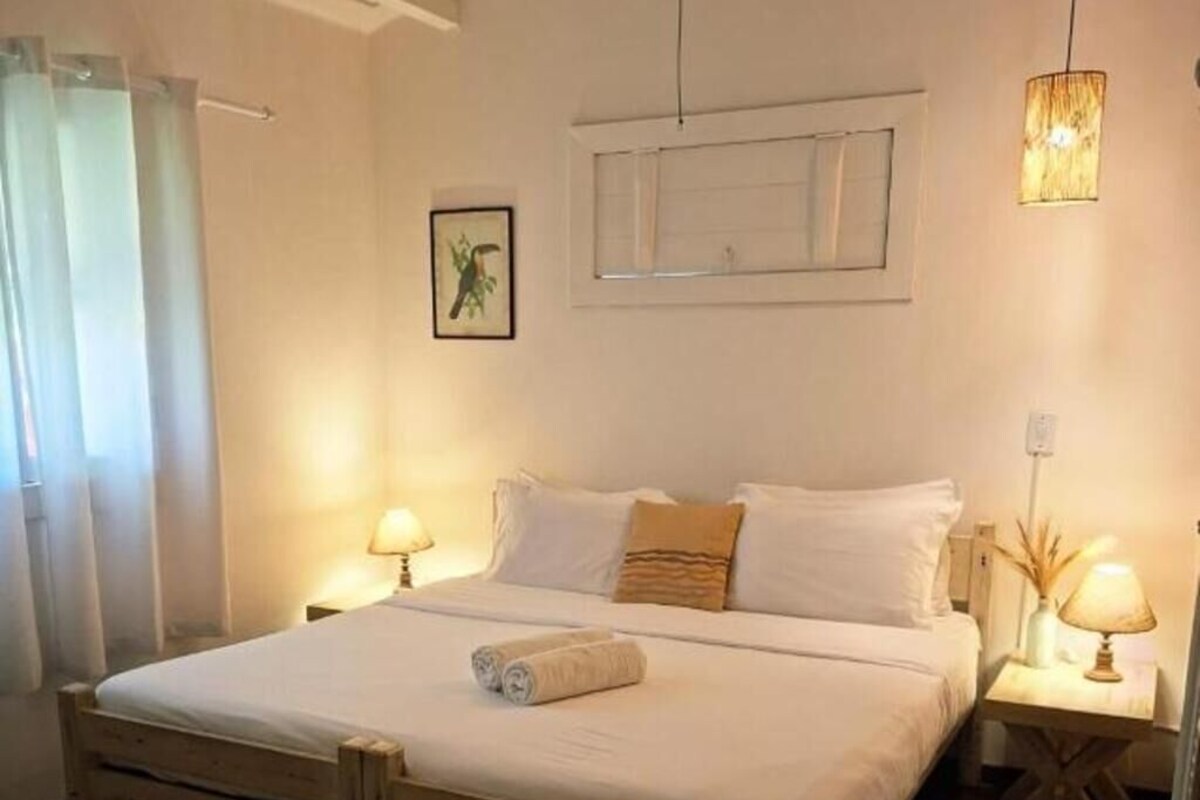 A bedroom with a wooden bed, two bedside tables with lamps, a window with curtains, and a wall-mounted picture.
