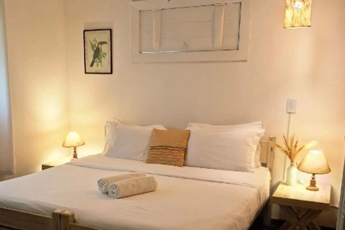 A hotel room with a bed, two bedside lamps, a window with blinds, and a framed picture on the wall.