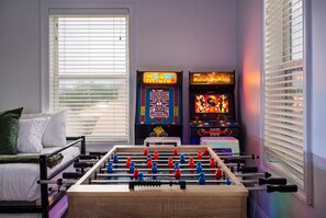 Una stanza con un tavolo da calcio, due giochi arcade, un letto con cuscini e una finestra con persiane.