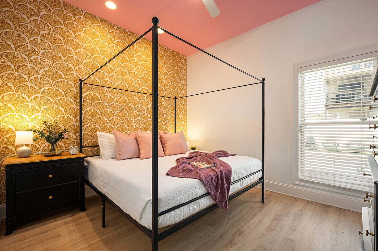 A four-poster bed with a white and pink bedding set, a bedside table with a lamp, and a wall with a leaf pattern.
