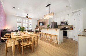 A modern kitchen with white cabinets, a central island with stools, a dining table with chairs, and a ceiling fan. - MilSViewsI PoolI BarrelSaunaIGulf ViewsI25Steps2BeachIGame RoomIPCB Beachlife (Panama City Beach)