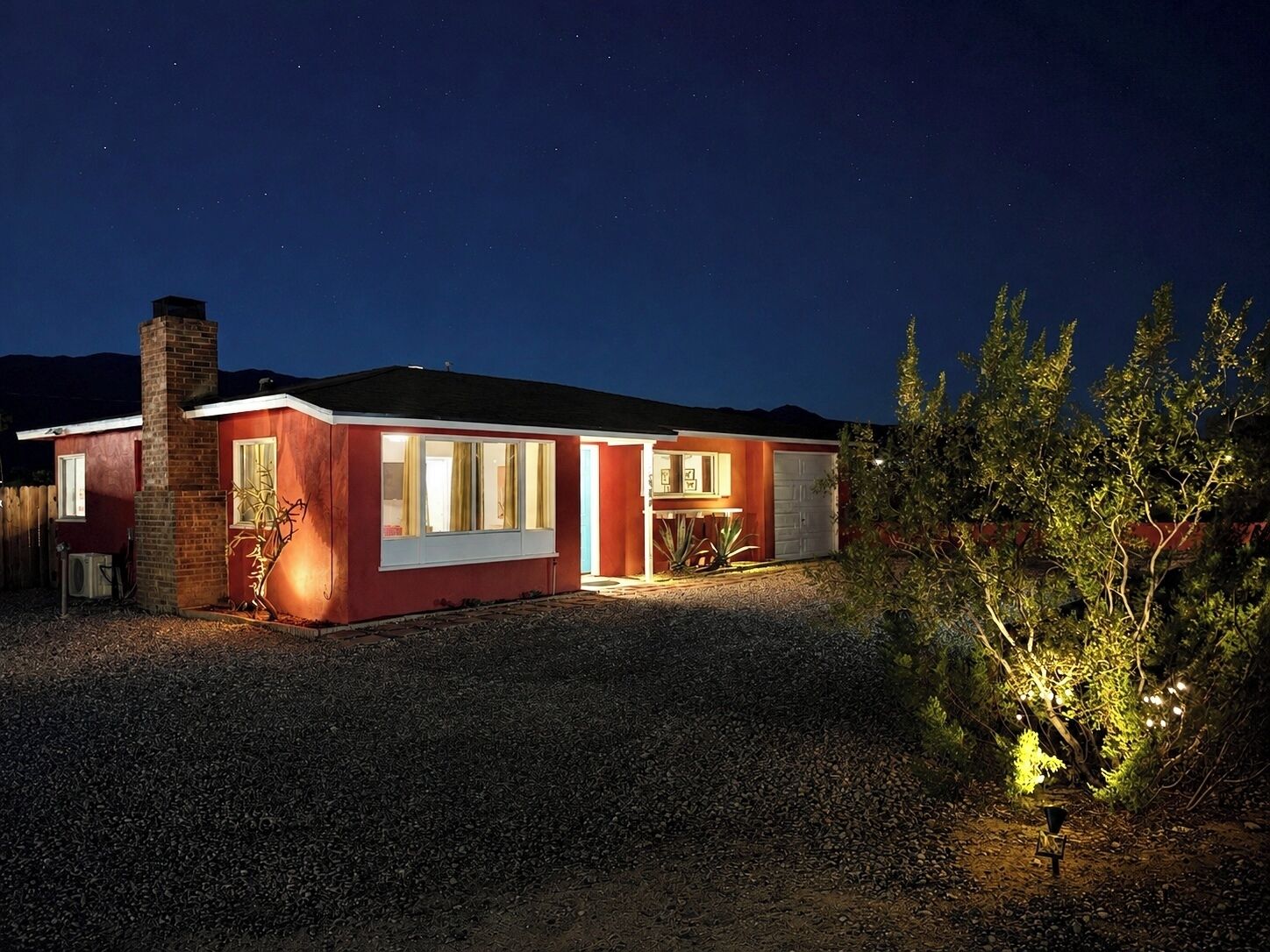 A single-story house with a red exterior and a chimney, surrounded by gravel and a small tree.