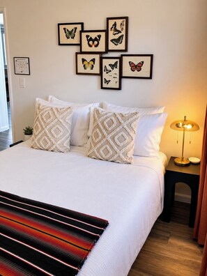 A neatly made bed with patterned pillows, a framed butterfly collection on the wall, and a bedside lamp.
