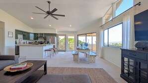 A spacious living area with a sofa, coffee table, and a dining table with chairs. There is a kitchen area with a ceiling fan and a large window providing a view.