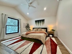 Ein Schlafzimmer mit einem Bett, einem Fenster mit Vorhängen, einem Deckenventilator, einem Nachttisch mit einer Lampe und einem Wandbild.