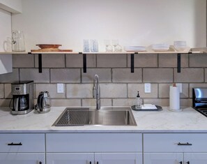 A kitchen with a stainless steel sink, a coffee maker, and a kettle. There are shelves above the sink holding bowls and glasses.