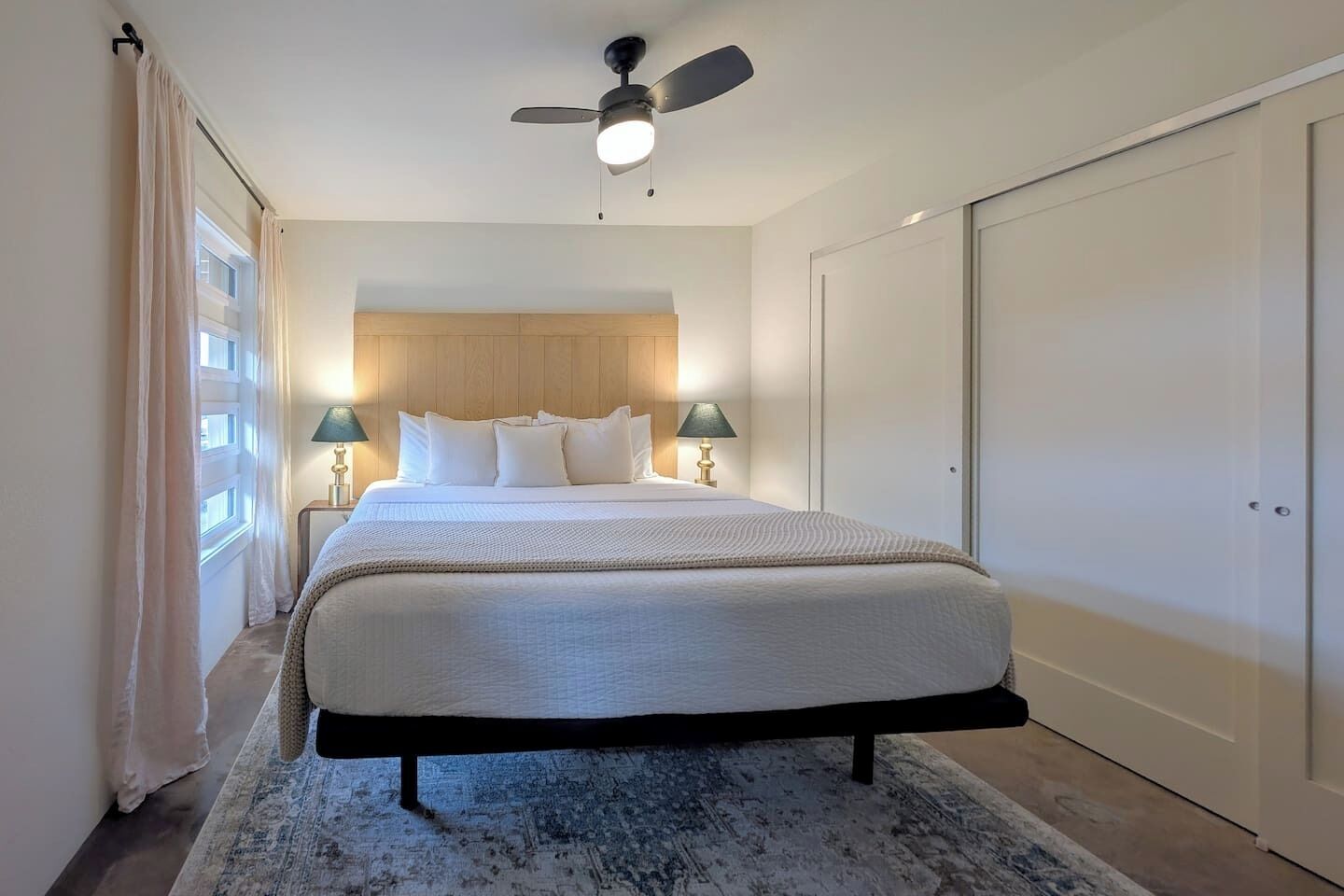 Une chambre à coucher avec un lit, deux lampes de chevet, un ventilateur de plafond et une grande fenêtre avec des rideaux.