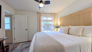 A bedroom with a bed, a ceiling fan, a window with curtains, a bedside lamp, and a wooden headboard. - Hale Maluhia, Newly renovated, 1BR, sleeps 3. (Waikoloa Village)