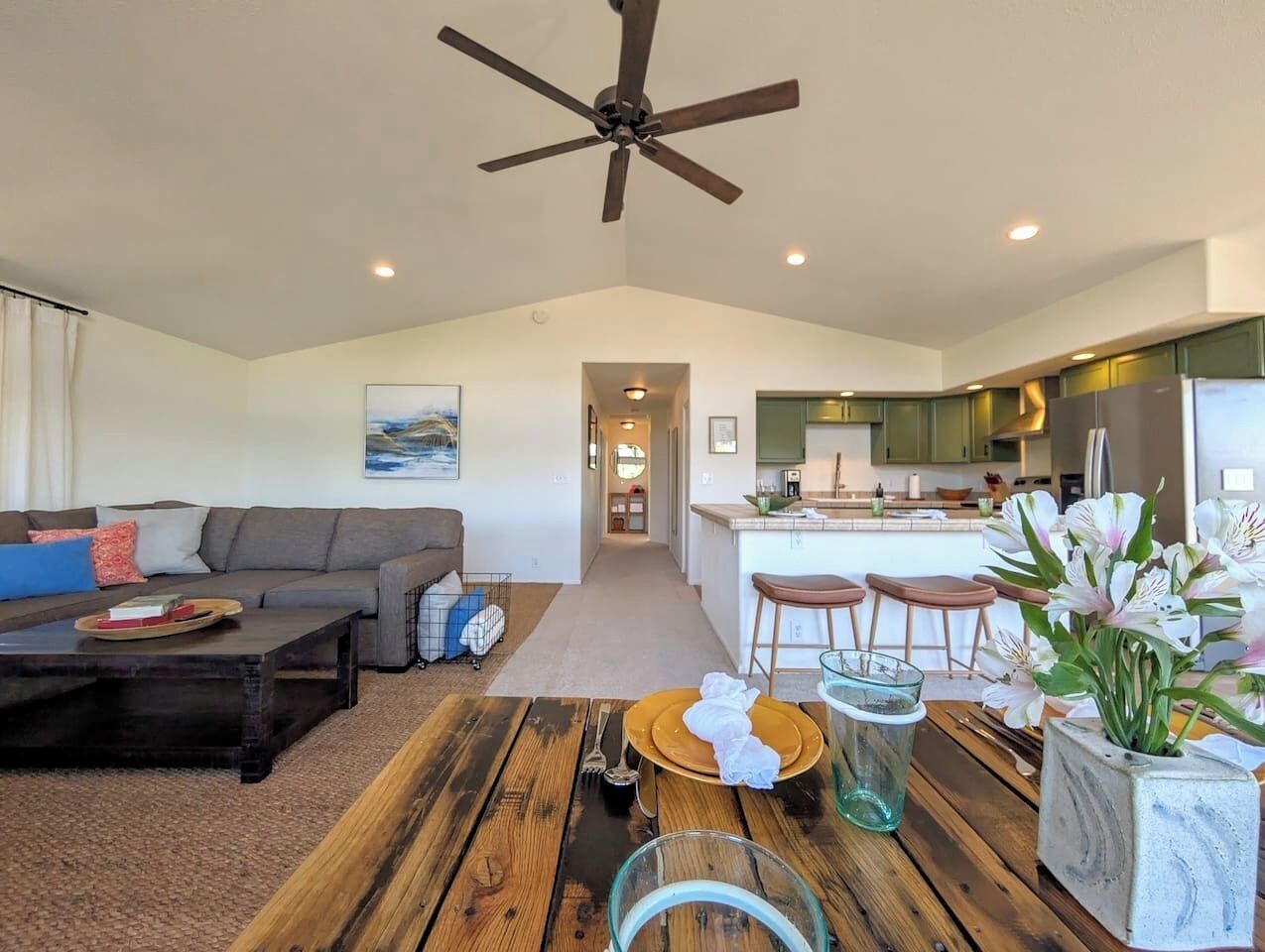 A modern living room with a sofa, coffee table, and dining table. There is a kitchen area with bar stools and a refrigerator. A ceiling fan is visible, and the room has a high ceiling.