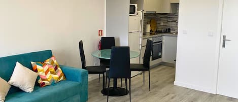 A modern living room with a teal sofa, a dining table with chairs, and a kitchen area with appliances.