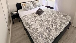 Un dormitorio con cama, mesita de noche, una obra de arte en la pared y una ventana con cortinas.