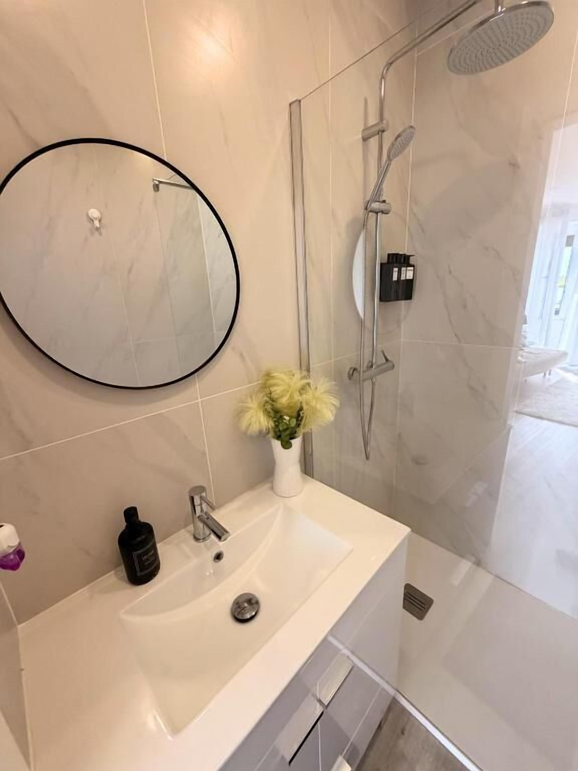 Un bany amb un lavabo blanc, un mirall rodó, una cabina de dutxa de vidre i un ram de flors.
