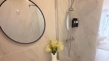 Une salle de bain avec un lavabo blanc, un miroir rond, une cabine de douche en verre et un vase avec des fleurs.