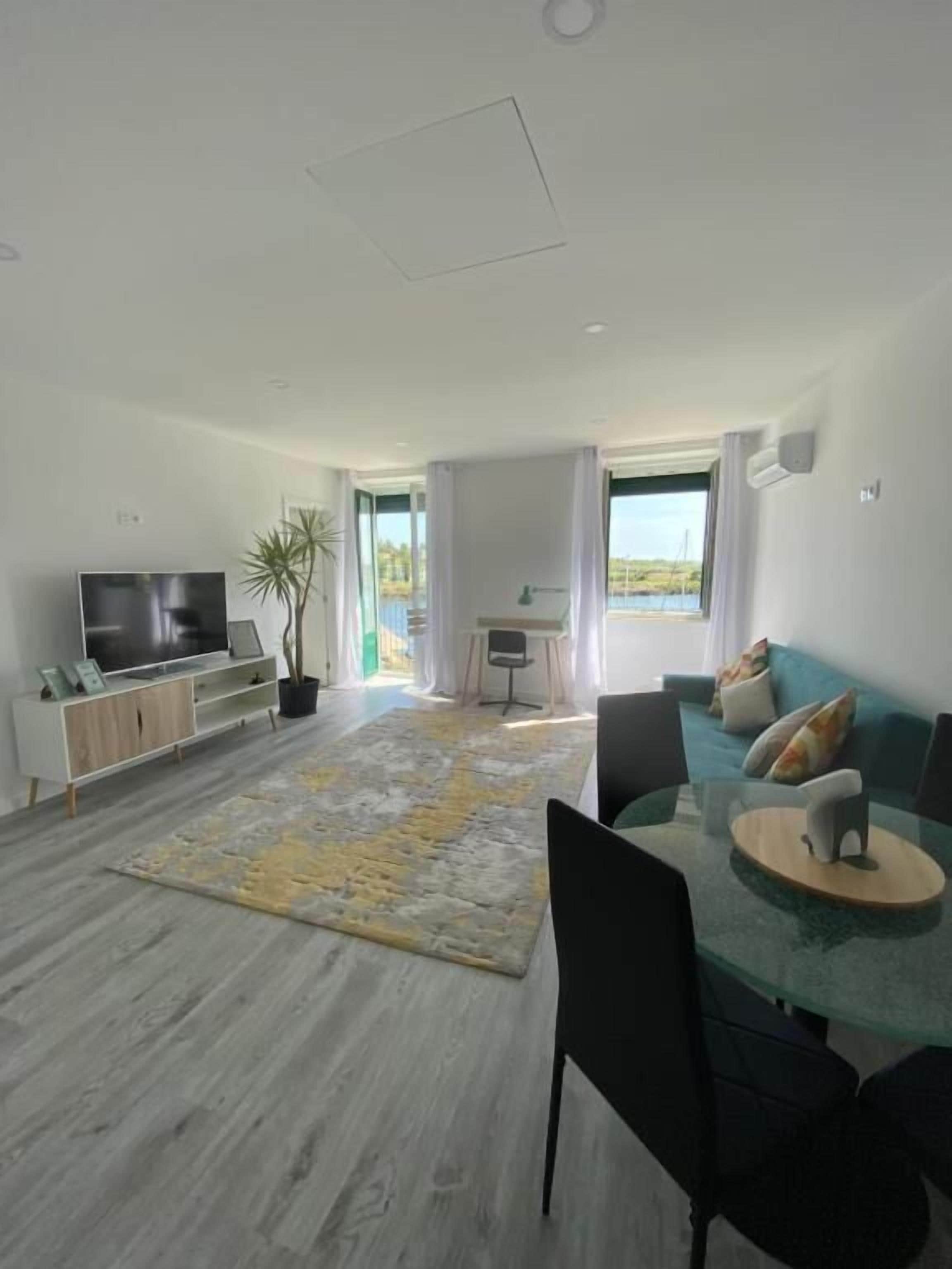 A modern living room with a flat-screen TV, a dining table with chairs, a sofa, and a large window offering a view of the outdoors.