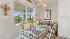 Área de refeições com uma mesa comprida, cadeiras e vista para o exterior. Há uma cruz na parede e um quadro emoldurado sobre a mesa.