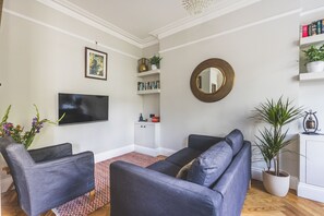 Een woonkamer met een flatscreen televisie, twee fauteuils, een kamerkleed met dessin, een boekenkast, een spiegel en potplanten.