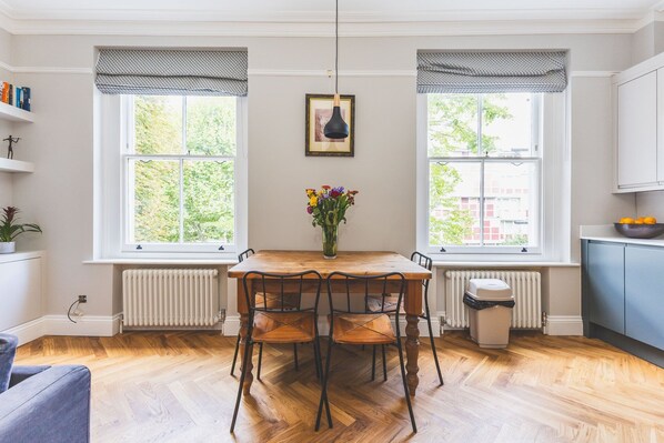 Een zithoek met een houten tafel en stoelen, twee ramen met zonwering, een radiator en een lijst met een afbeelding aan de muur.
