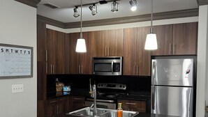 A modern kitchen with wooden cabinets, stainless steel appliances, a black countertop, and two white bar stools.