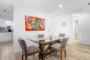 A modern dining area with a glass-top table, four upholstered chairs, and a colorful abstract painting on the wall. - Spacious 3BR Hollywood Home, Backyard & BBQ, Close to Beach & Hard Rock (Hollywood)