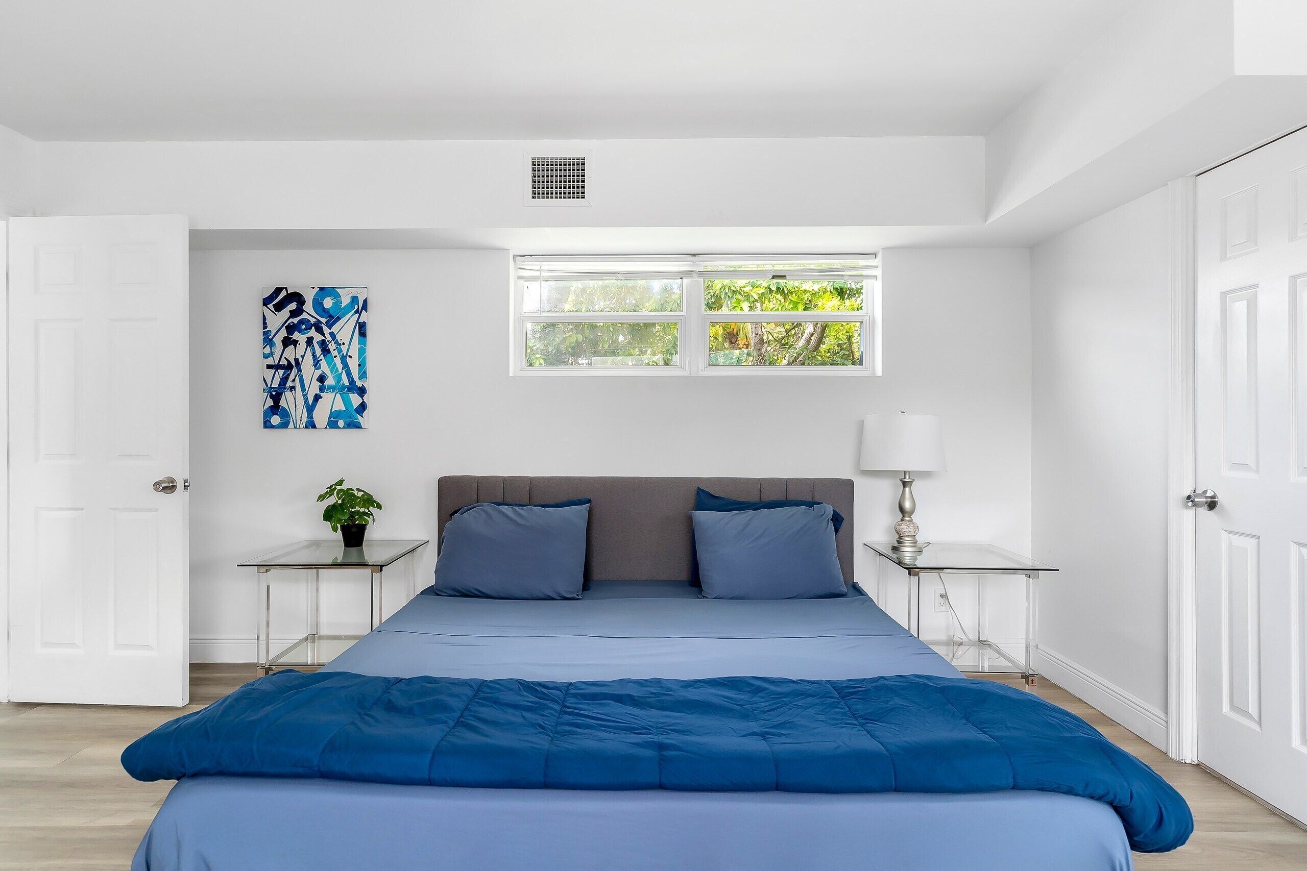 Un dormitorio con una cama grande, un ventanal con vistas a zonas verdes, una mesita de noche con una lámpara y una obra de arte colgada en la pared.