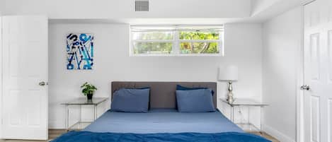 Un dormitorio con una cama grande, un ventanal con vistas a zonas verdes, una mesita de noche con una lámpara y una obra de arte colgada en la pared.