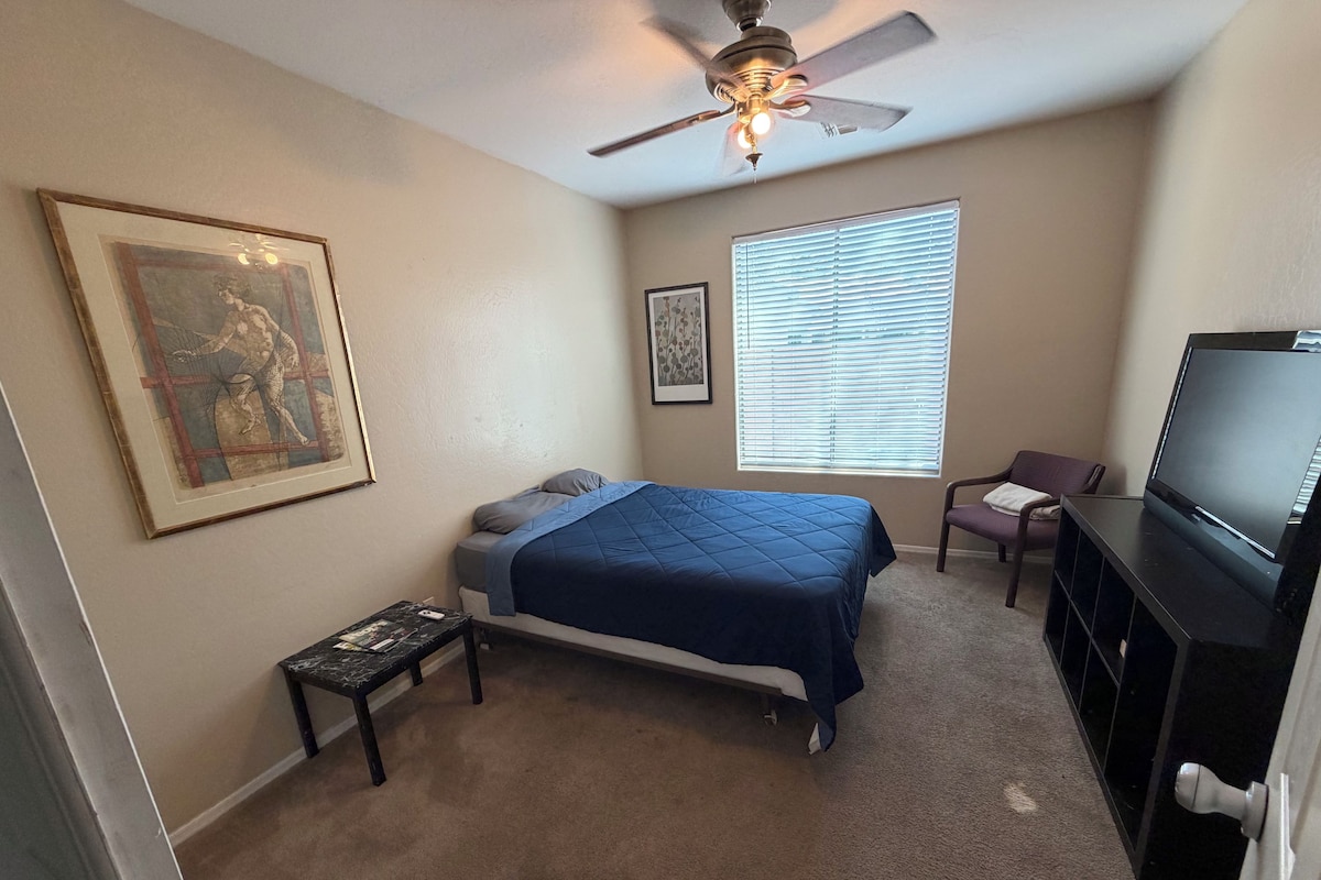 A bedroom with a bed, a ceiling fan, a television, a window with blinds, and a framed picture on the wall.