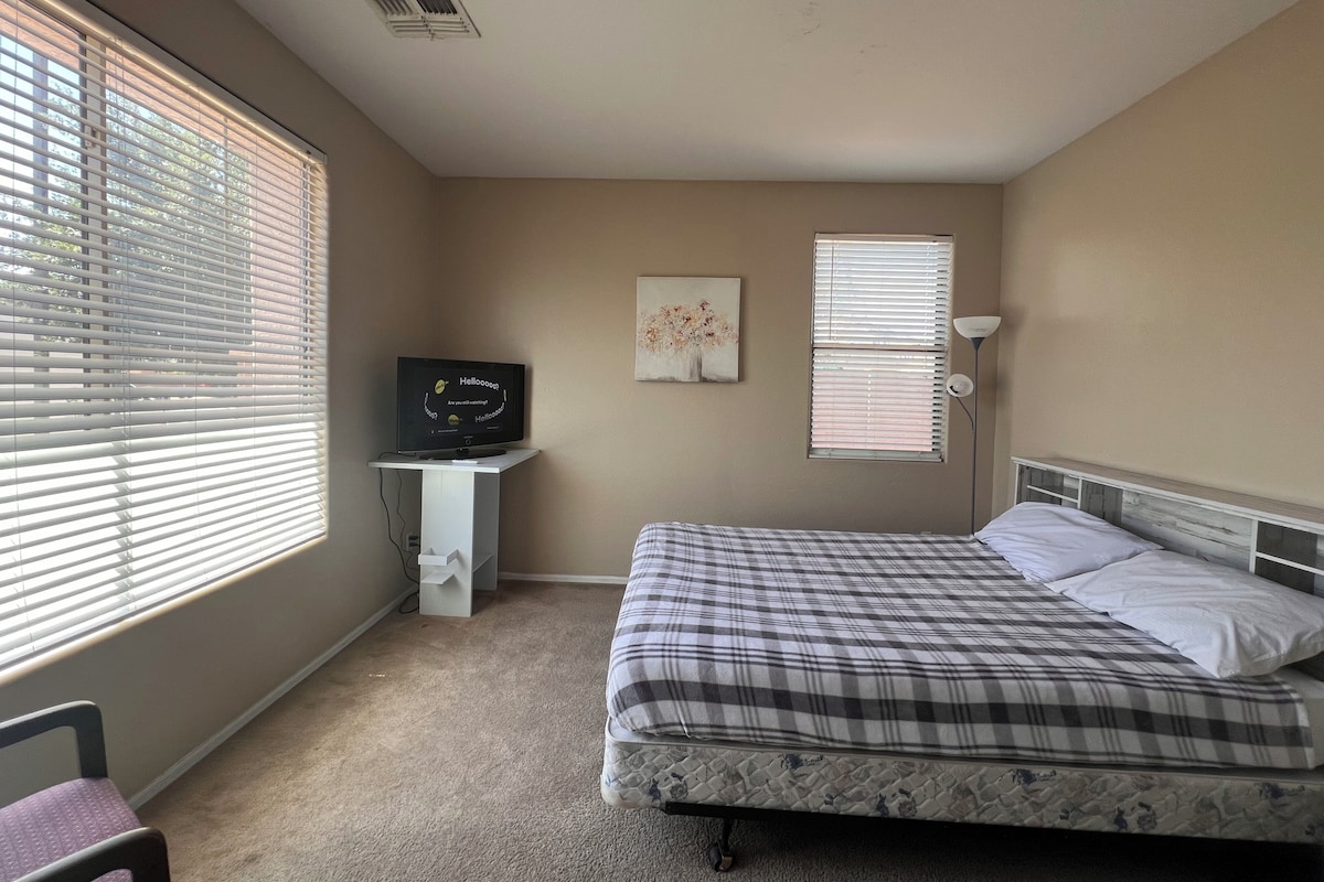 A bedroom with a bed, a desk with a television, a window with blinds, and a lamp.