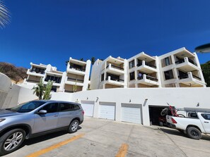 Un edificio residencial de varios pisos con balcones, un estacionamiento con una camioneta SUV plateada y una camioneta pickup blanca.