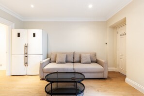 A living room with a beige sofa, a glass coffee table, and a white refrigerator. - Pimlico 2-Bed | Victoria & Belgravia (London)