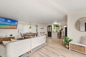 A modern living room with a sofa, dining area, kitchen, and a large TV. - Kaneohe Bay Hale (Kaneohe)