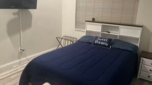 A bedroom with a bed, a bedside table, a nightstand, a window with blinds, and a wall-mounted TV.