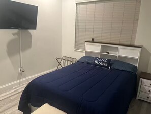 A bedroom with a bed, a bedside table, a nightstand, a window with blinds, and a wall-mounted TV.