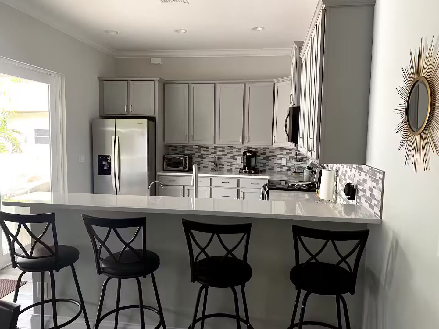Una cocina moderna con armarios blancos, un refrigerador de acero inoxidable, un microondas y una cafetera. Hay cuatro taburetes negros en la isla.