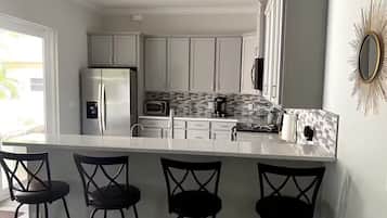 Una cocina moderna con armarios blancos, un refrigerador de acero inoxidable, un microondas y una cafetera. Hay cuatro taburetes negros en la isla.