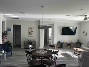 A modern living room with a dining area, a flat-screen TV mounted on the wall, and a ceiling fan.